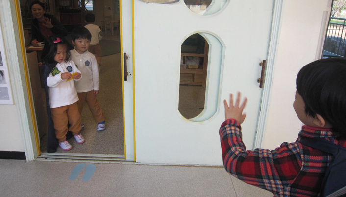 가방을 메고 복도에 서 있는 아이가 교실 안의 두 명의 친구를 향해 웃이며 손을 흔들고 있는 모습이다