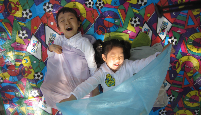 돗자리 위에 누워 있는 두 아이가 각각 연보라색, 하늘색 천을 잡고 웃고 있다