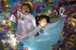 돗자리 위에 누워 있는 두 아이가 각각 연보라색, 하늘색 천을 잡고 웃고 있다