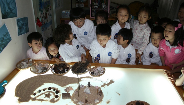 라이트박스 위에 놓여진 모래와 나뭇가지, 종이 등을 바라보고 있는 아이들의 모습이다