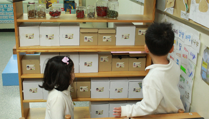 교실 안에서 두 아이가 메시지 박스에 붙어있는 아이들의 사진을 보고 있는 모습이다