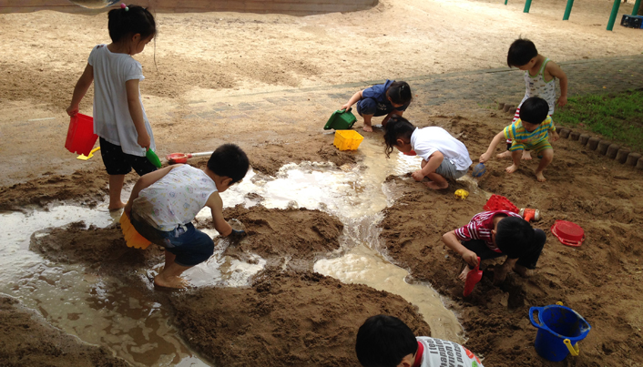 물이 흐르고 있는 모래사장 위에서 아이들이 삽과 통을 들고 모래놀이를 하고 있는 모습이다