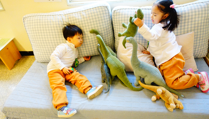 소파에 앉아 공룡인형을 갖고 놀고 있는 남자아이와 여자아이의 모습이다