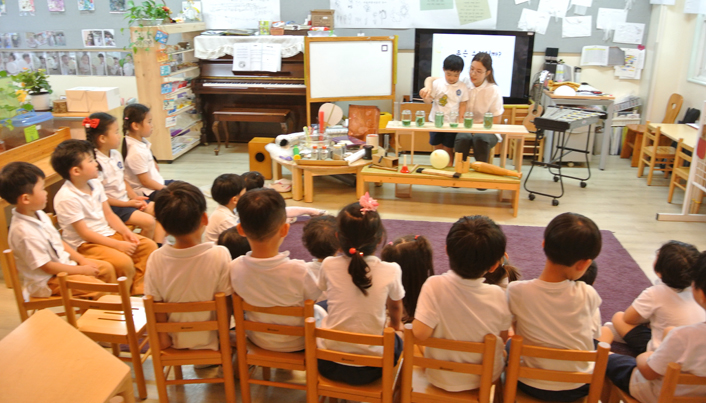 아이들이 의자에 앉아 교실 앞에서 선생님과 한 친구가 실험을 하는 모습을 지켜보고 있다