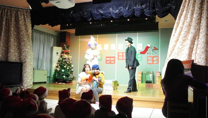 무대 위에서 네 명의 선생님이 연극을 하고 있고 바닥에는 산타모자를 쓴 아이들이 앉아 연극을 보고 있다